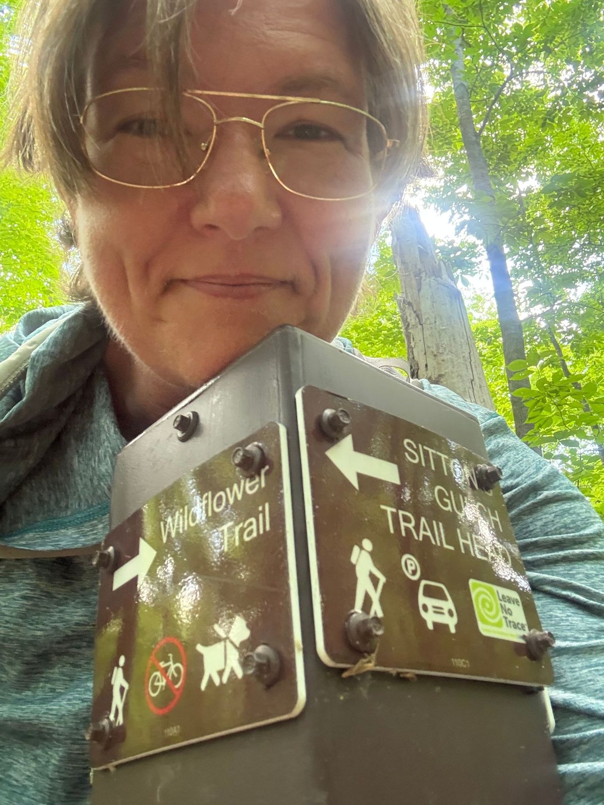 RJ Kirkland outdoors on a wooded trail, standing beside a trailhead signpost.